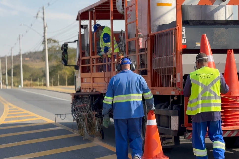 Trabalhadores da equipe de sinalização viária acompanham o caminhão de pintura de faixas das vias enquanto sinalizam o trabalho com cones.