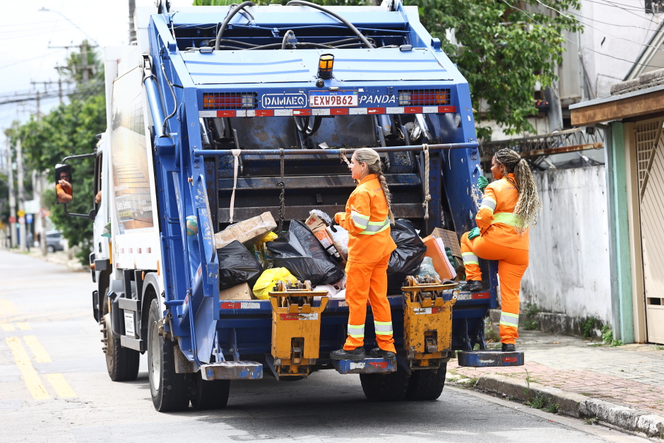 Duas coletoras uniformizadas com os trajes laranjas da Urbam acompanham o caminhão da coleta na parte traseira do veículo