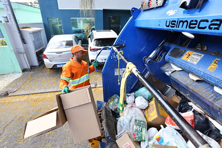 Coletor da Urbam vestido de laranja segura na parte traseira do caminhão de lixo enquanto realiza a coleta seletiva