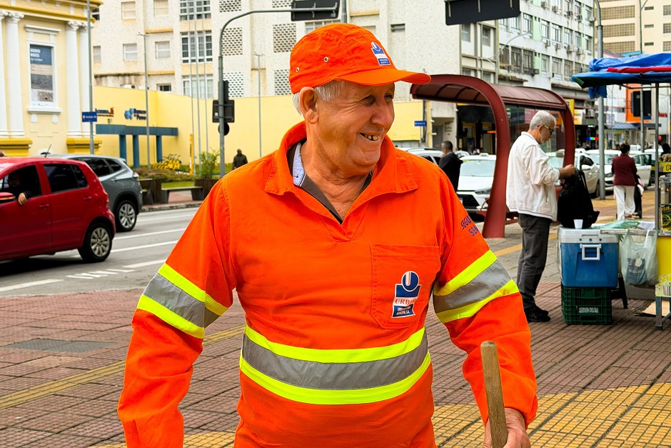 Seu Nelson aparece uniformizado em seu traje laranja da Urbam sorrindo enquanto varre as ruas do centro de São José